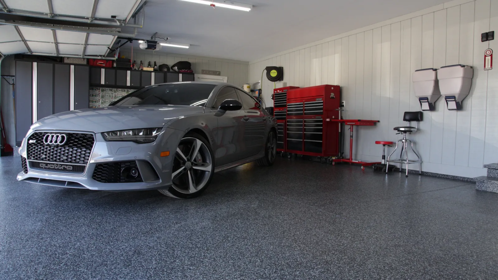 Silver Audi parked inside a clean, organized garage with red toolboxes and stools against white paneled walls.