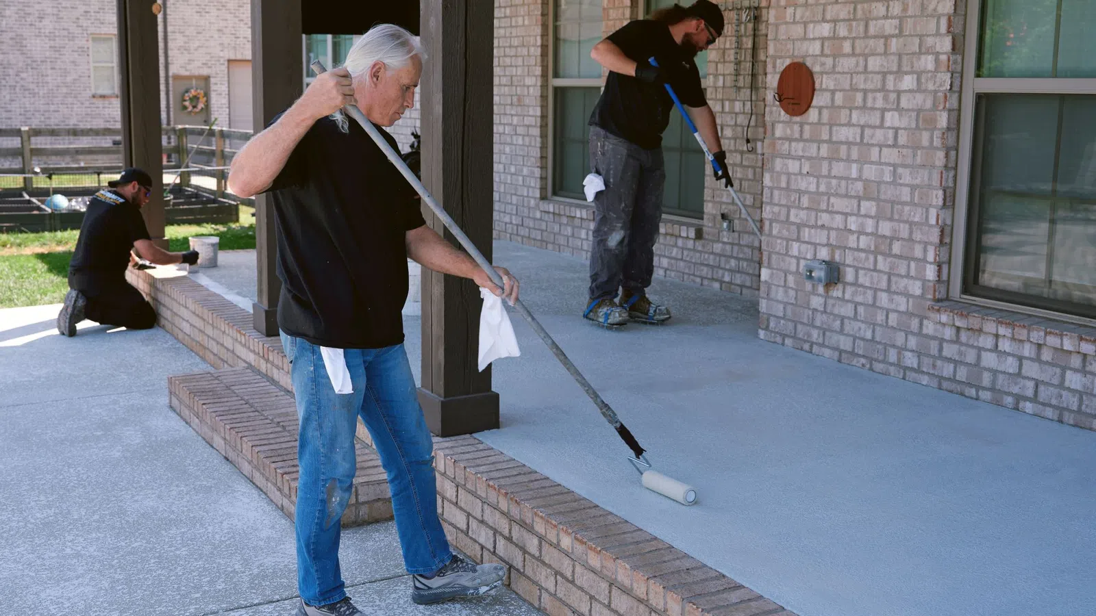 Three men painting a concrete porch grey outside a brick house under daylight with paint rollers and brushes.