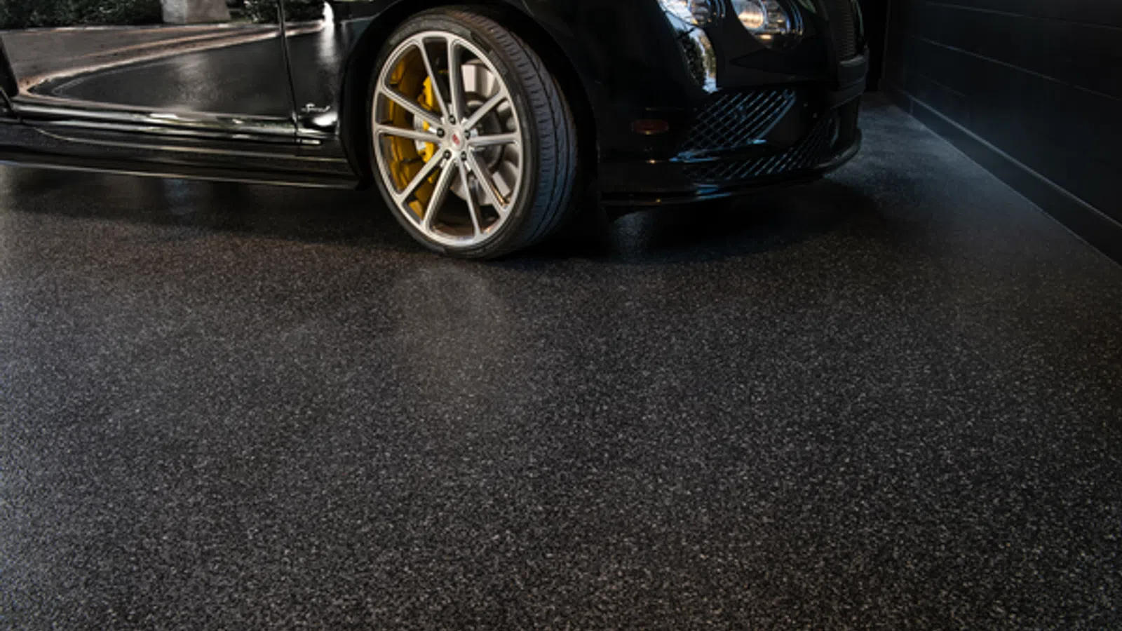 Close-up of a luxury black car wheel with yellow brake calipers on a polished dark speckled floor.