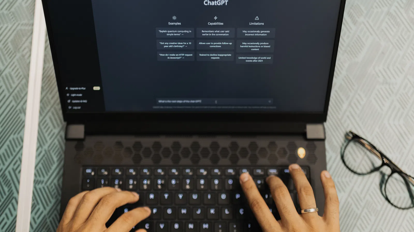 Person typing on a laptop displaying ChatGPT interface with eyeglasses on table beside it