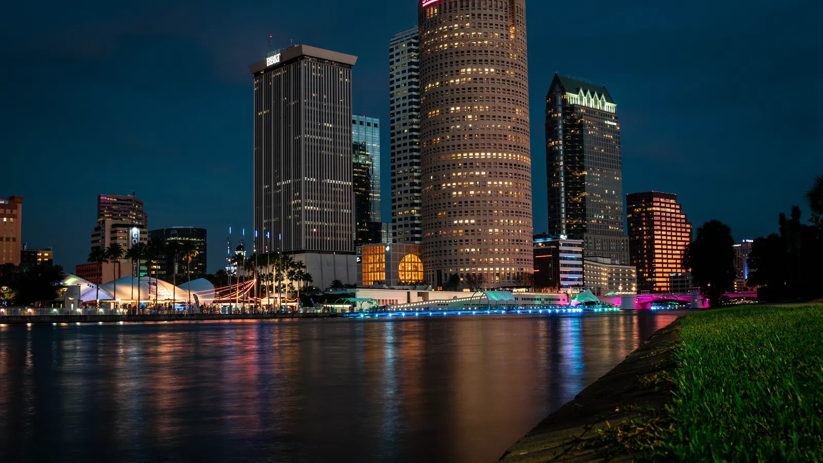 Nighttime city skyline with illuminated high-rise buildings reflecting on calm river water under a dark sky.