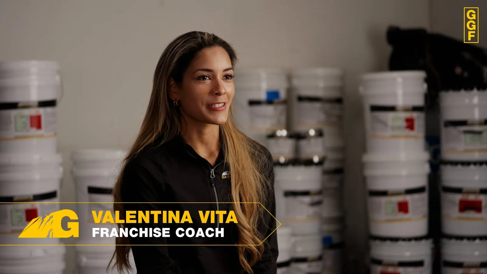 Franchise coach Valentina Vita speaking in front of stacked white buckets in a warehouse setting.