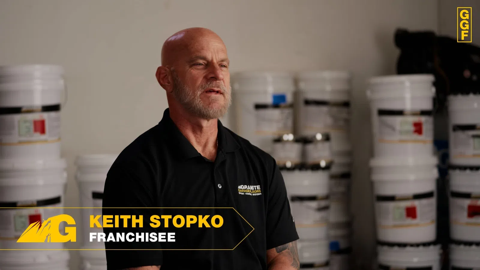 Man in black polo shirt with Agranite logo stands in front of stacked white buckets in warehouse setting.