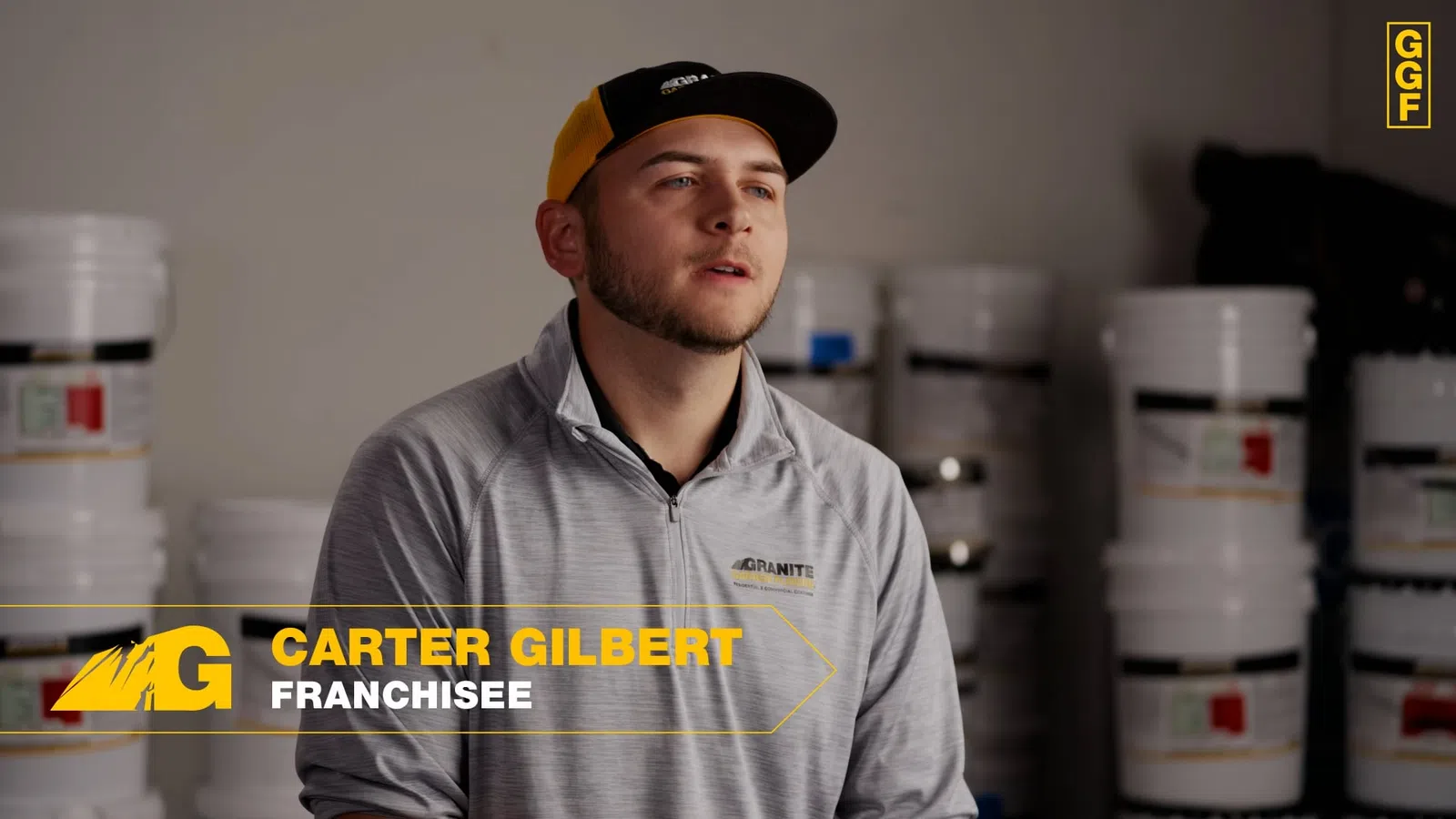 Young man in a gray Granite brand jacket and cap speaking in a warehouse with stacked buckets in background