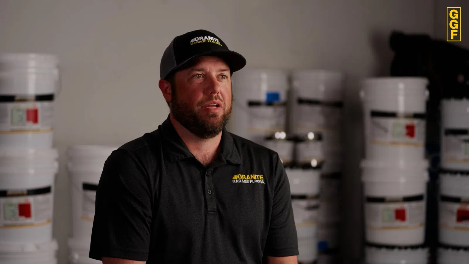 Man in Granite Garage Floors uniform and cap sitting indoors with stacked buckets of garage floor products in background.