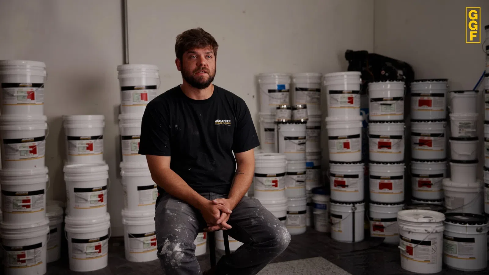 Man sitting on a stool in a warehouse surrounded by stacked white paint buckets.