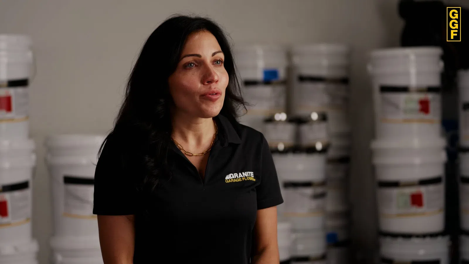 Woman wearing a Granite Garage Floors shirt stands in front of stacked white buckets in a warehouse setting.