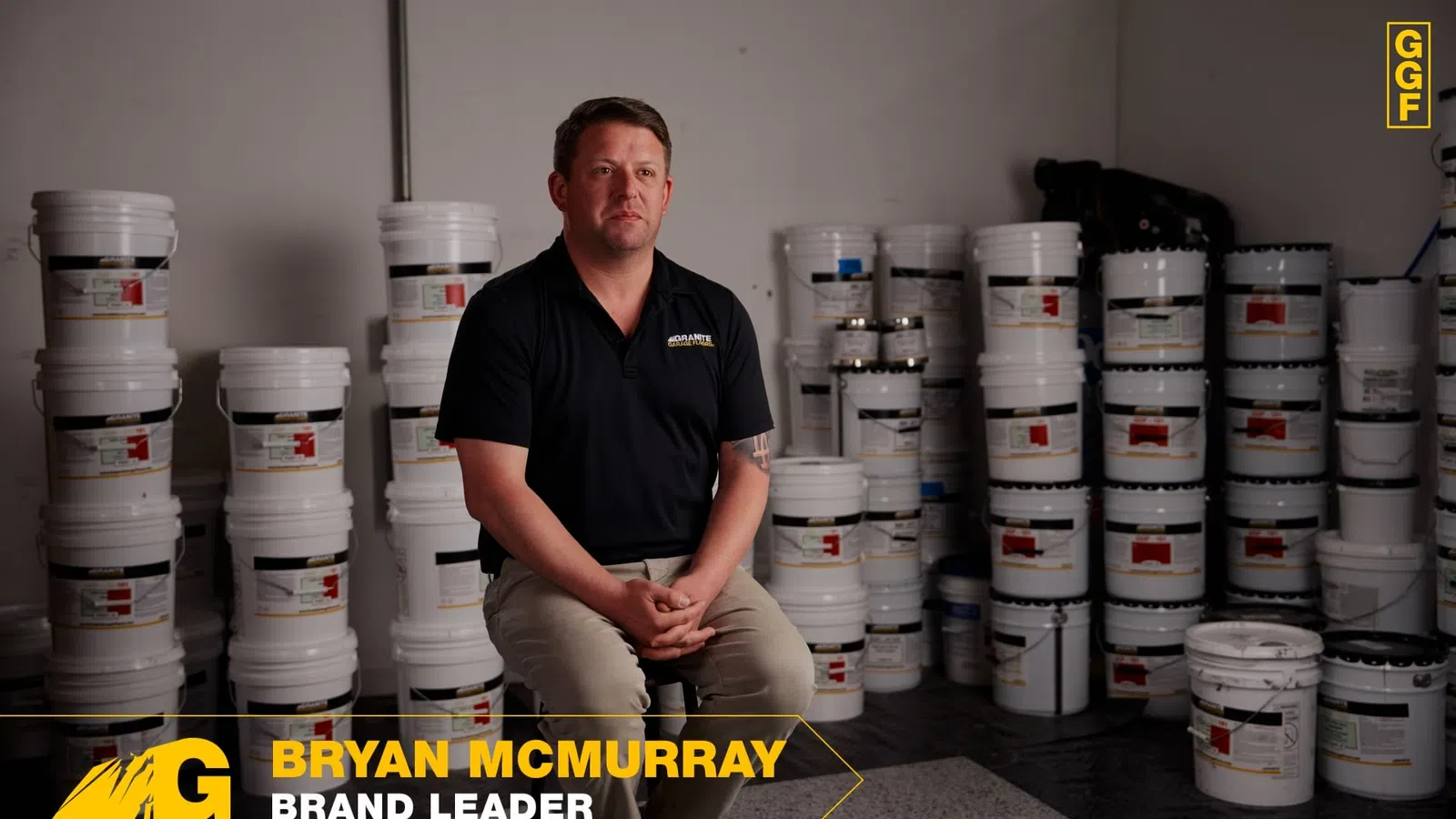Man in black shirt sitting surrounded by stacked white paint buckets in a storage room with gray walls.