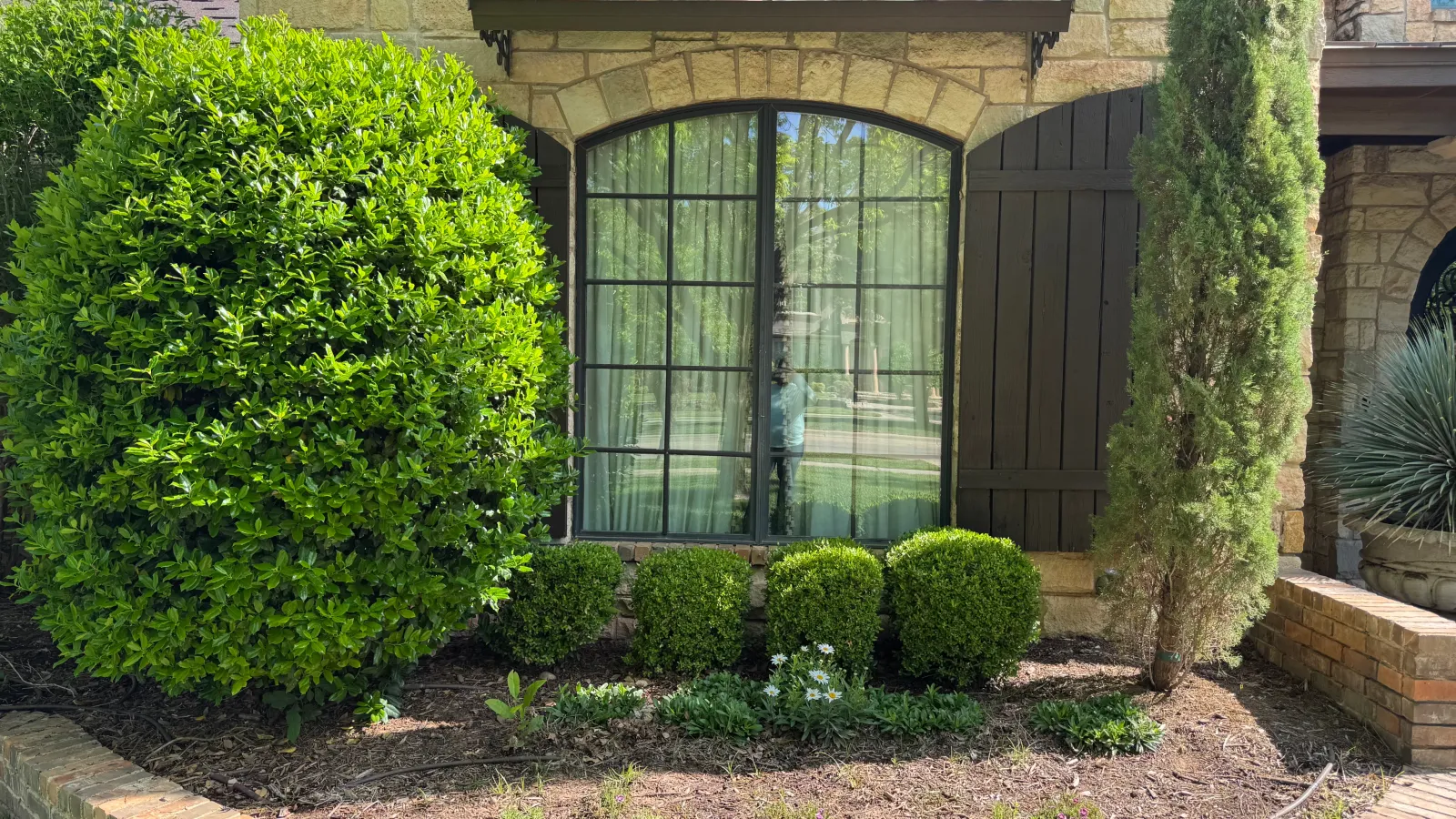 Stone house facade with arched window, black shutters, and neatly trimmed bushes in curved brick flower bed.