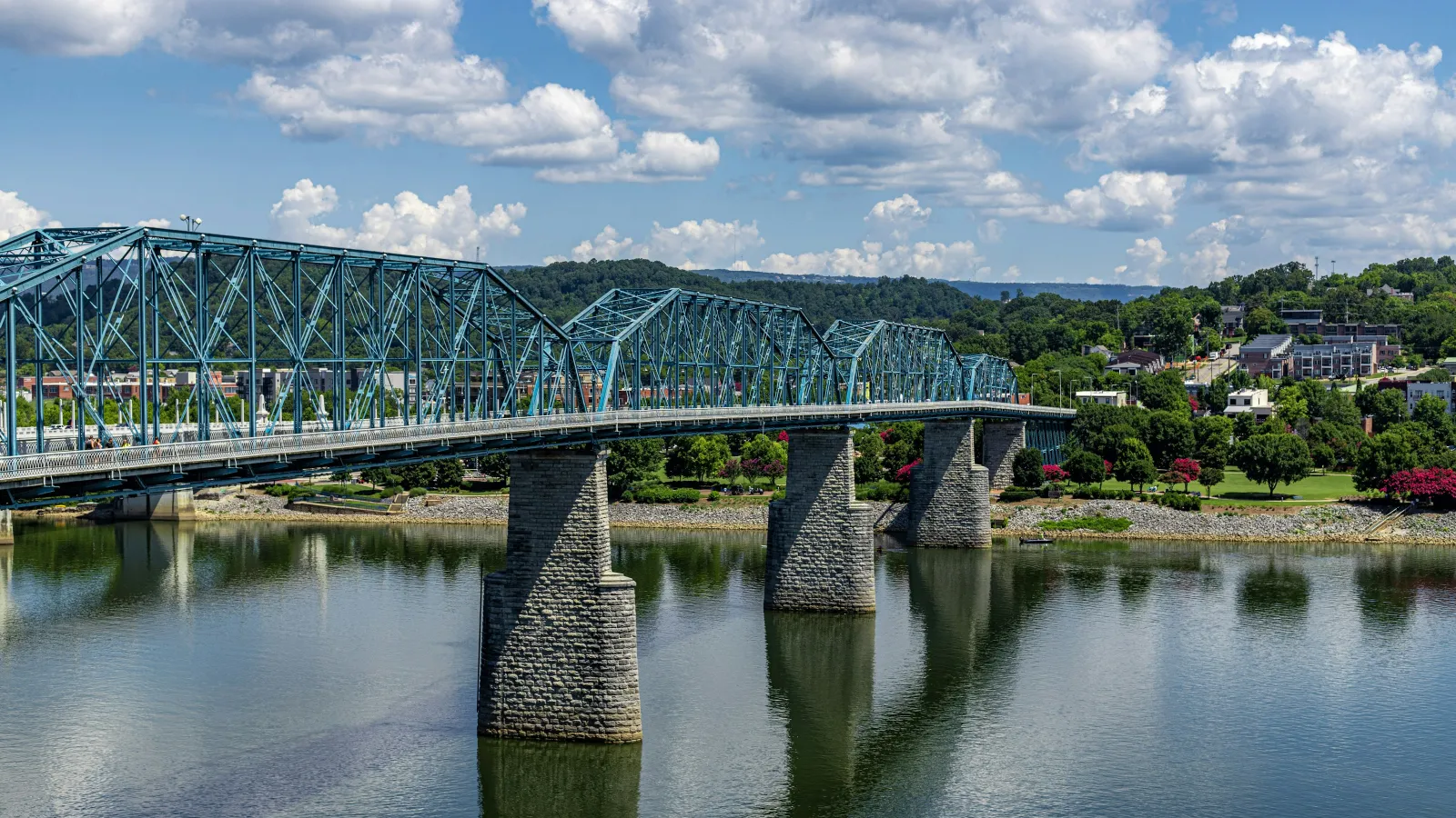 Blue steel truss bridge over calm river with stone piers under a partly cloudy sky in a green landscape