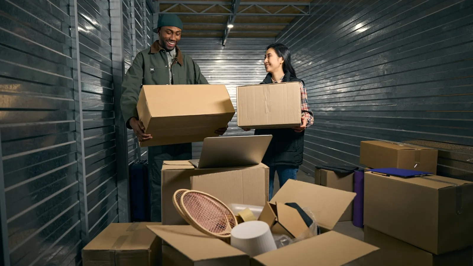 Two people smiling while unpacking boxes in a storage unit, surrounded by cardboard boxes.