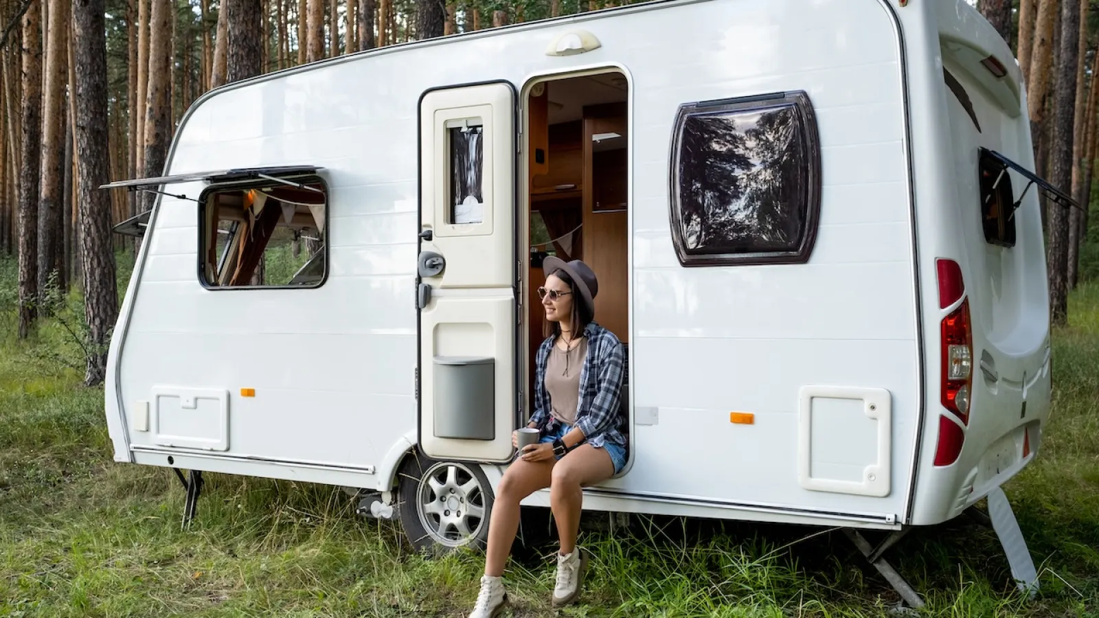 Woman enjoying nature beside a modern caravan in a forest setting.