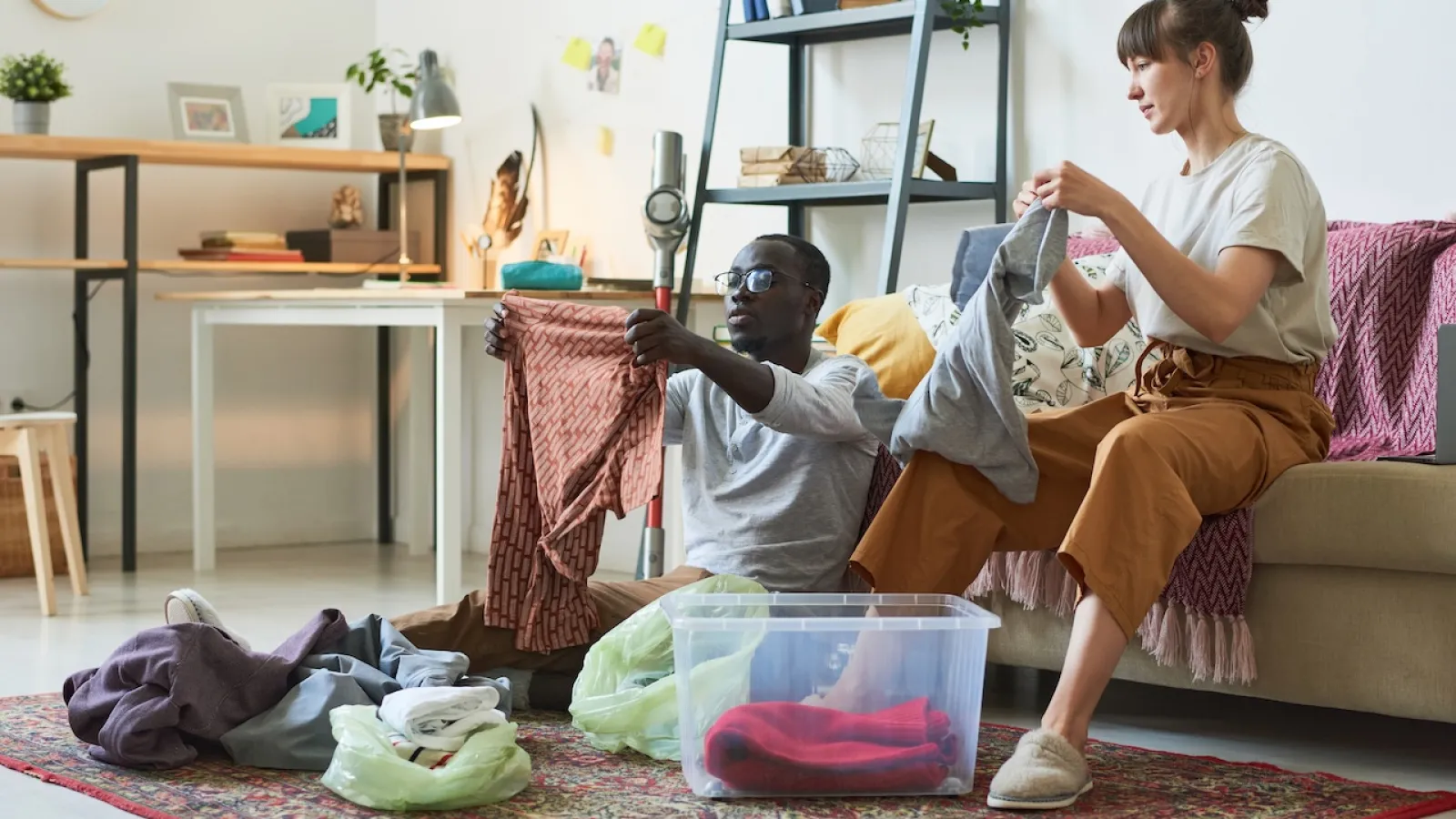 Couple sitting on floor and sofa sorting laundry in living room with clothes and plastic bins around.