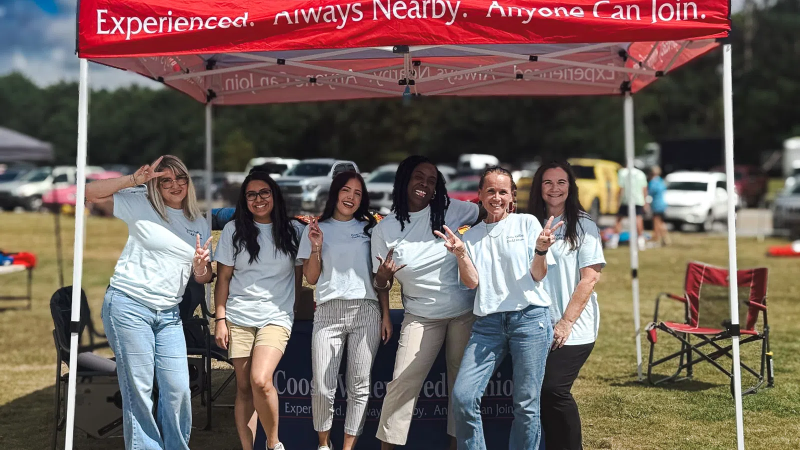 Group of six women posing and smiling under a Coosa Valley Credit Union event tent outdoors on a sunny day.