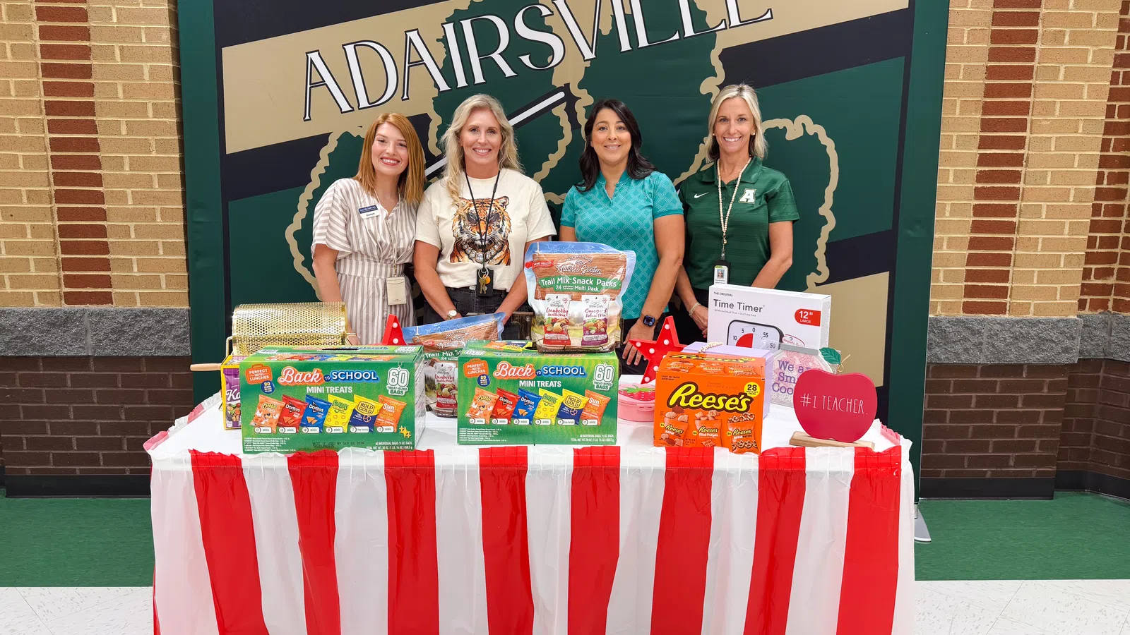 Four women standing behind a table with school supplies and snacks at Adairsville event with red and white striped tablecloth.