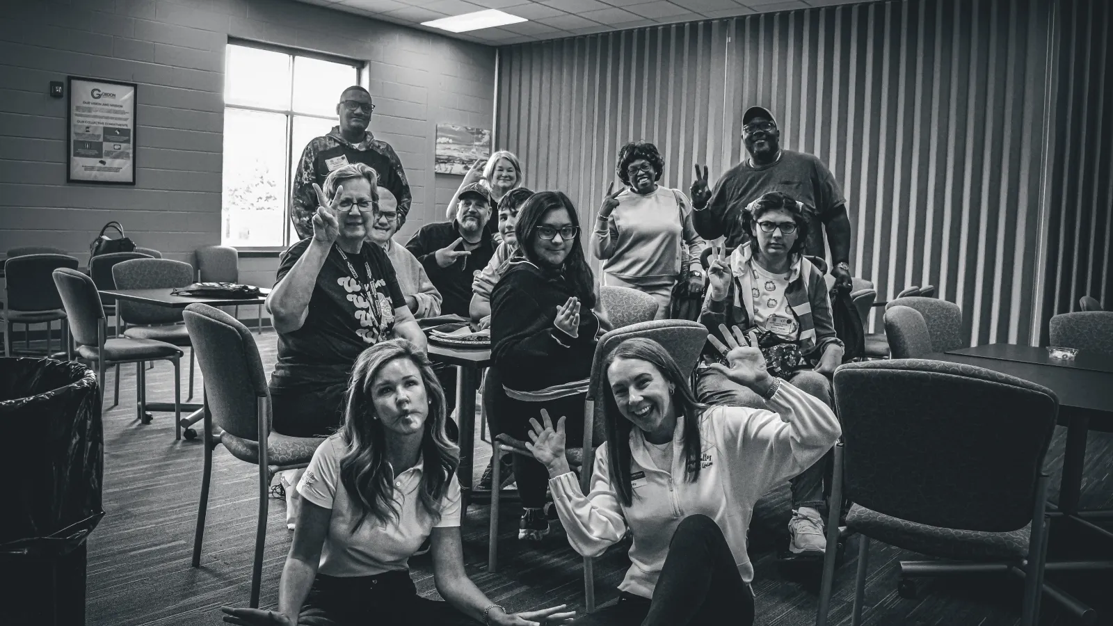 Black and white photo of diverse group posing playfully in a room with chairs and tables under fluorescent lights.