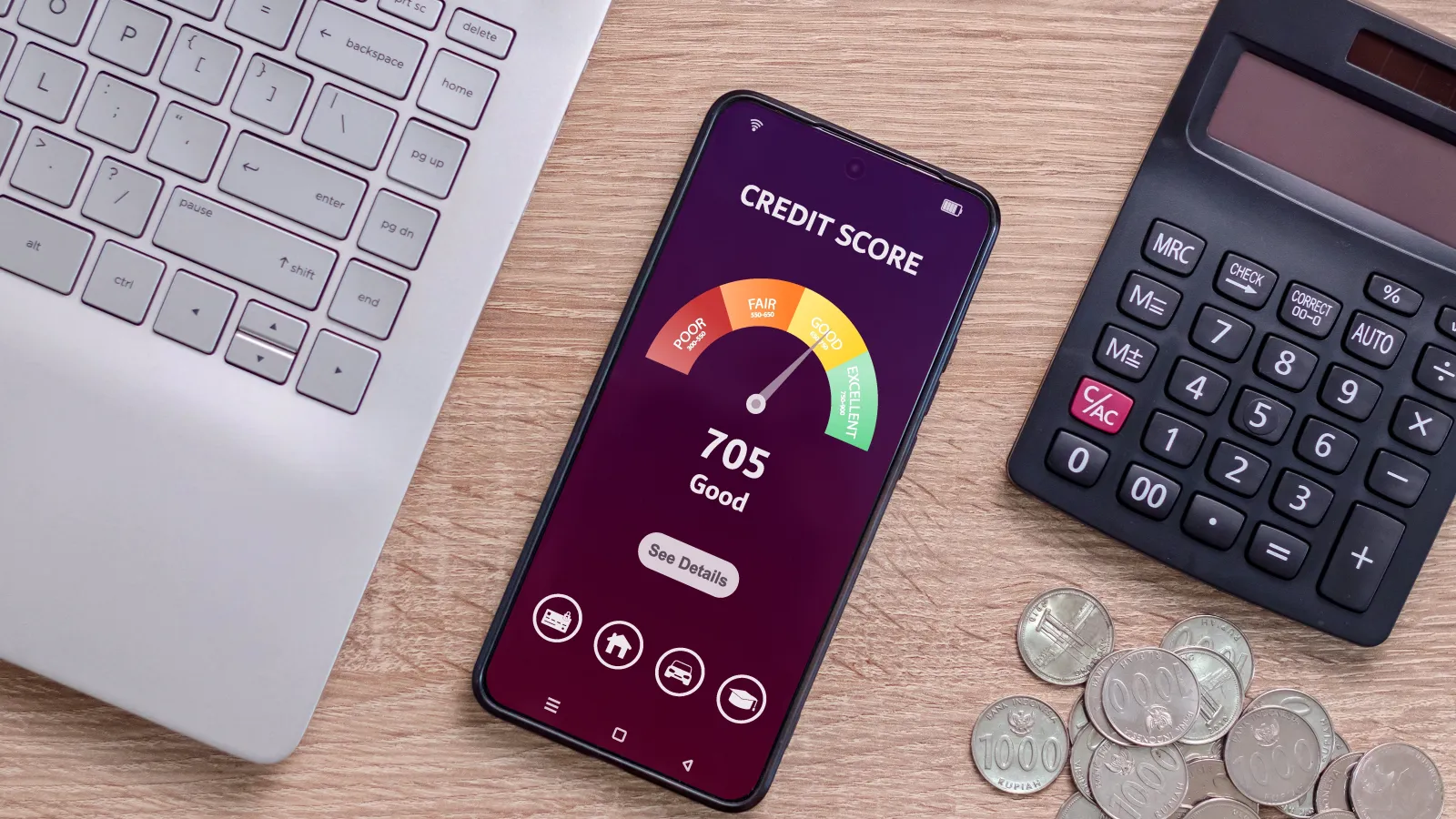 Smartphone showing a credit score of 705 labeled good, placed between a laptop, calculator, and coins on a wooden desk.