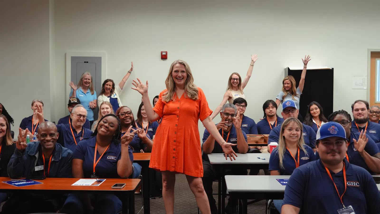 Smiling group of diverse students and teacher in orange dress posing happily in classroom with raised hands.