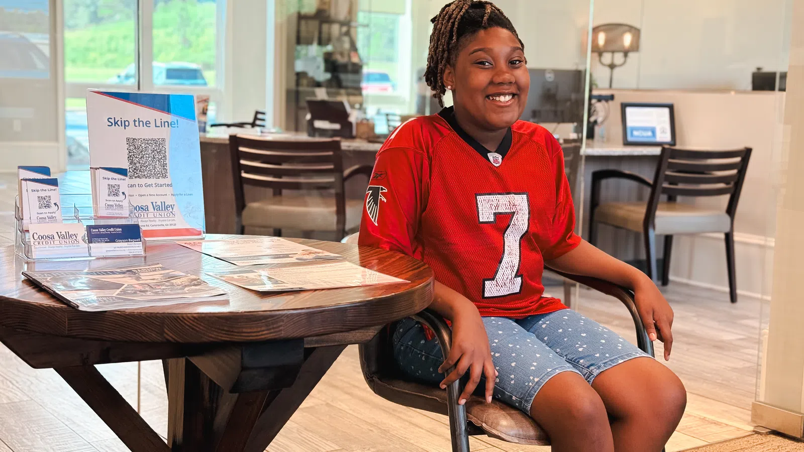 Smiling girl in a red jersey sitting in a modern bank lobby near informational brochures on a wooden table.