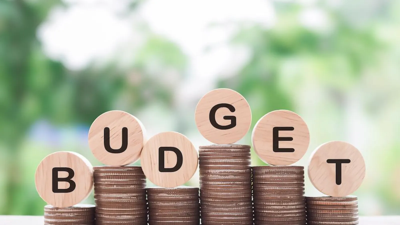 Stacked coins with wooden blocks spelling BUDGET on a blurred green background symbolizing financial planning.