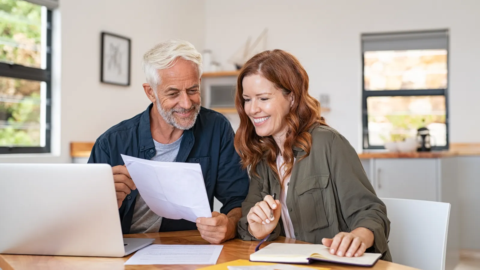 Mature couple reviewing documents and planning finances together at home with laptop and notebook