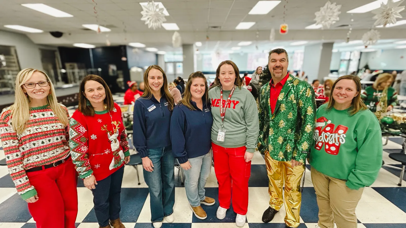 Seven people in festive holiday sweaters and attire posing indoors on a checkered floor with holiday decorations.