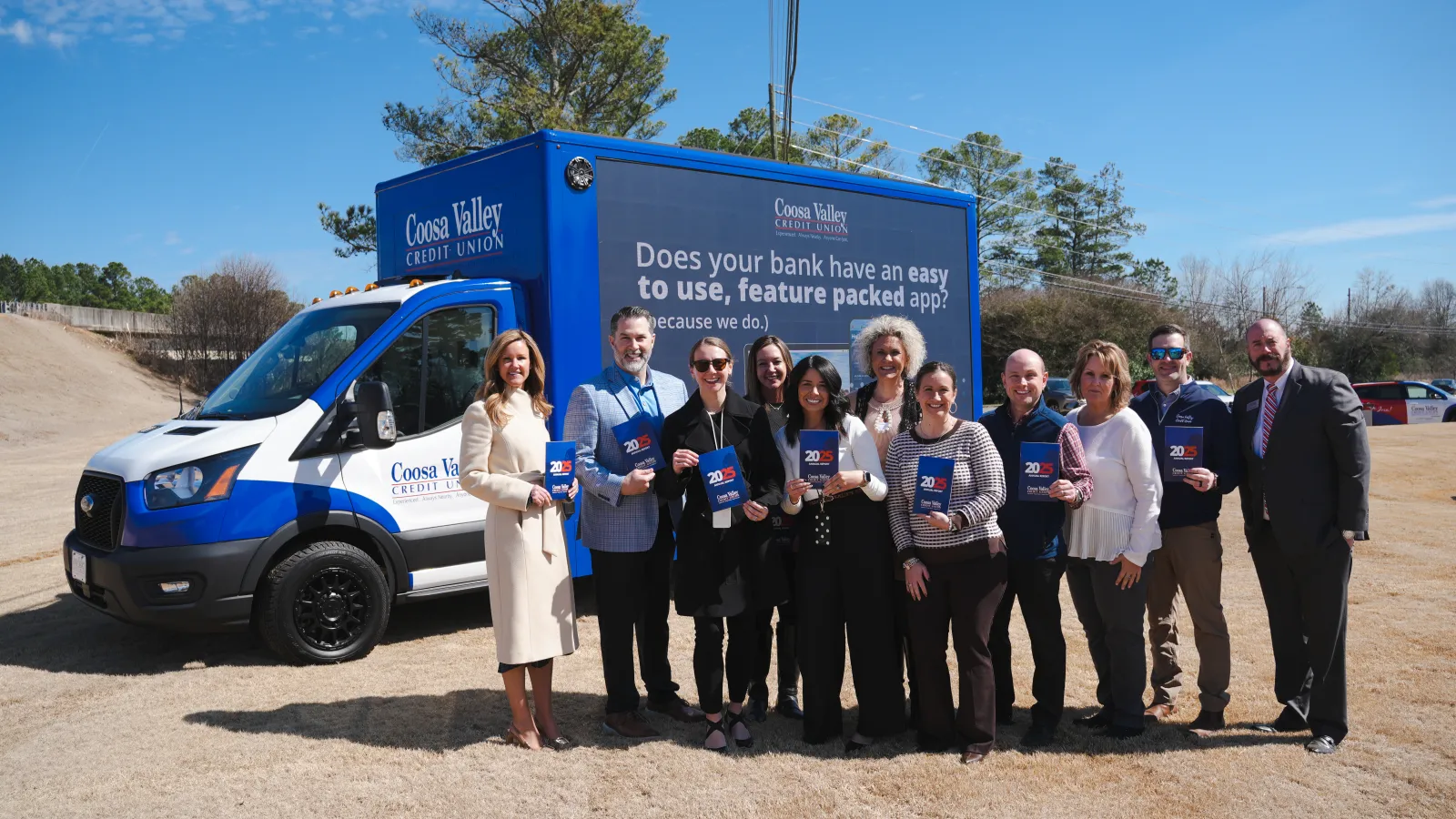 Group of people standing in front of a Coosa Valley Credit Union van holding brochures outdoors on a sunny day.