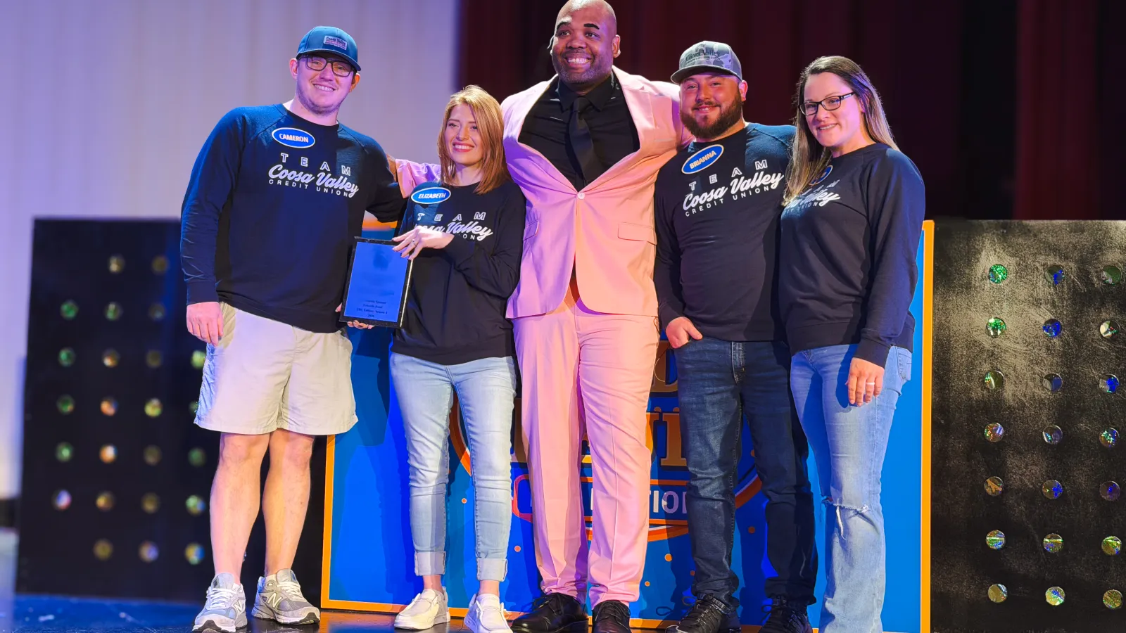 Five smiling people on stage, two holding a plaque, with one person in a pink suit and others in casual attire.