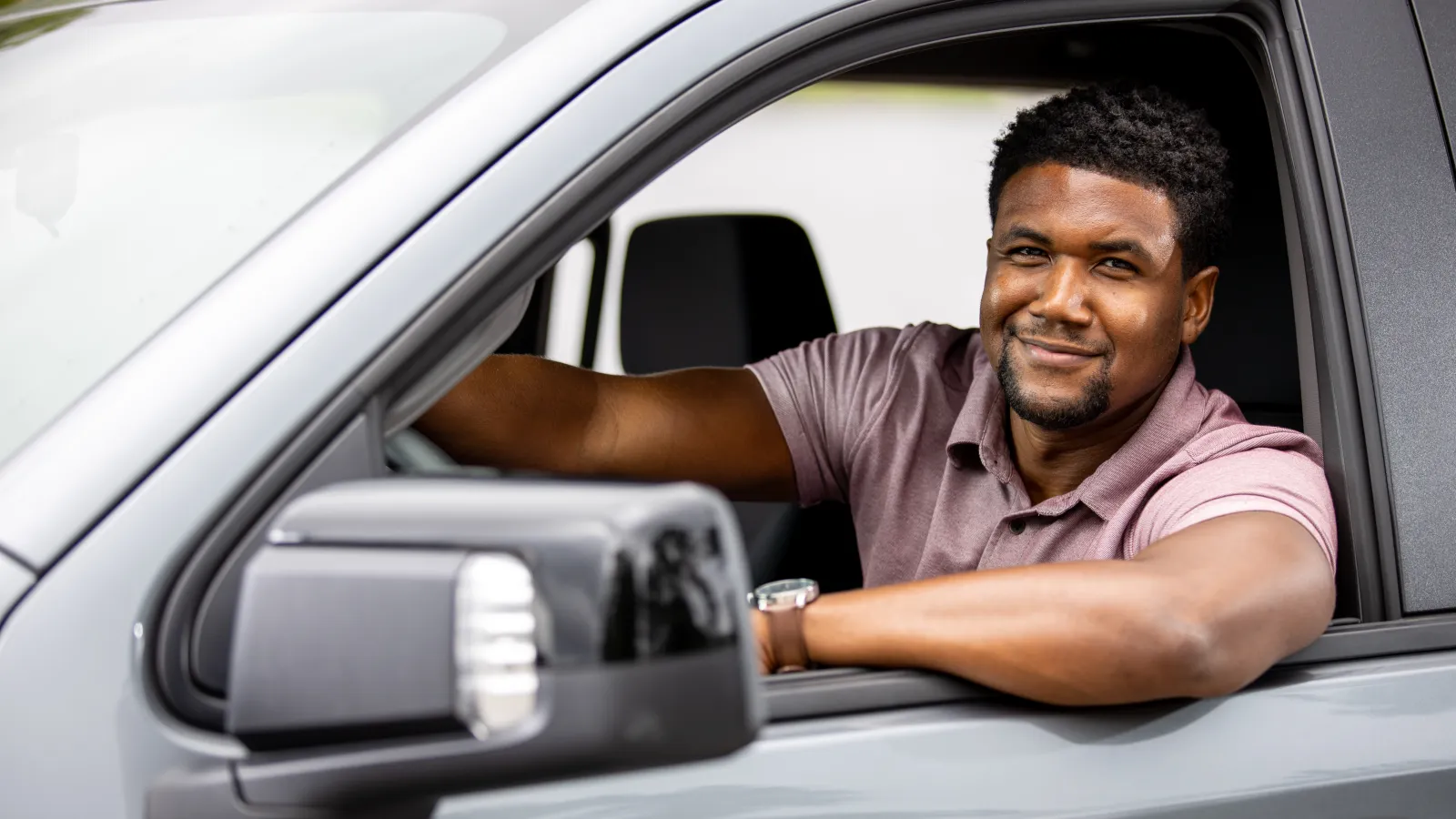 Smiling man sitting in a grey car with his arm resting on the window frame on a sunny day.