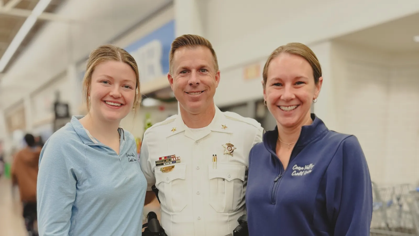Three smiling people posing indoors, including a uniformed sheriff and two casually dressed women in light and dark blue tops.