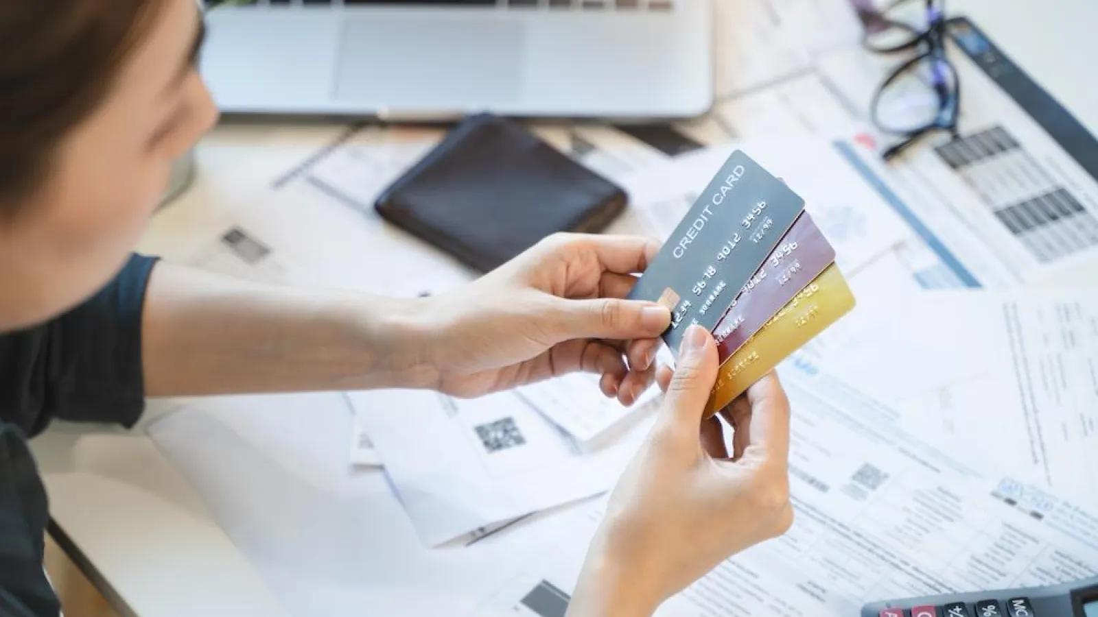 Person holding multiple credit cards over financial documents with a laptop and calculator in the background