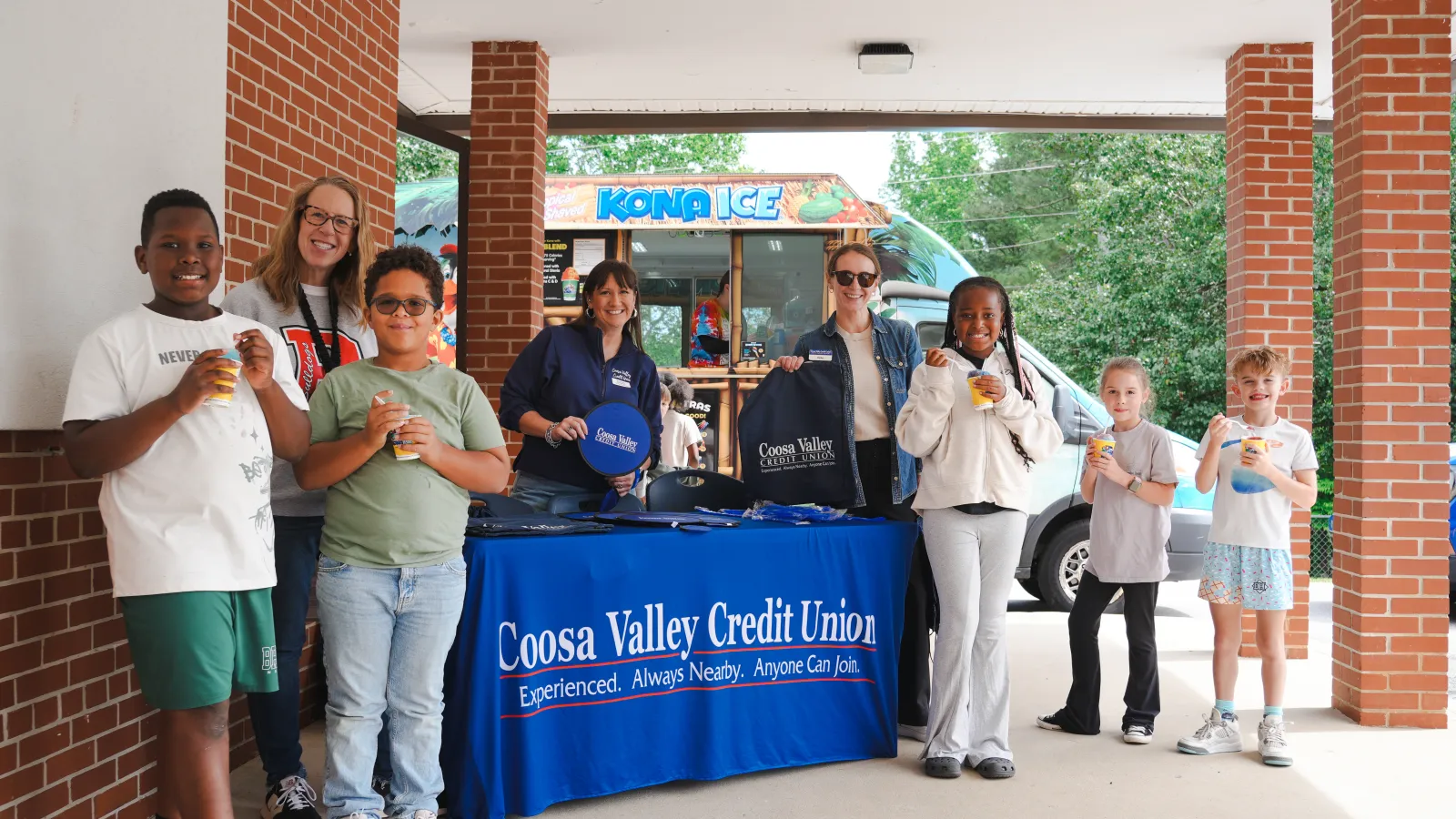 Group of children and adults enjoying drinks in front of Coosa Valley Credit Union booth and Kona Ice truck outdoors.
