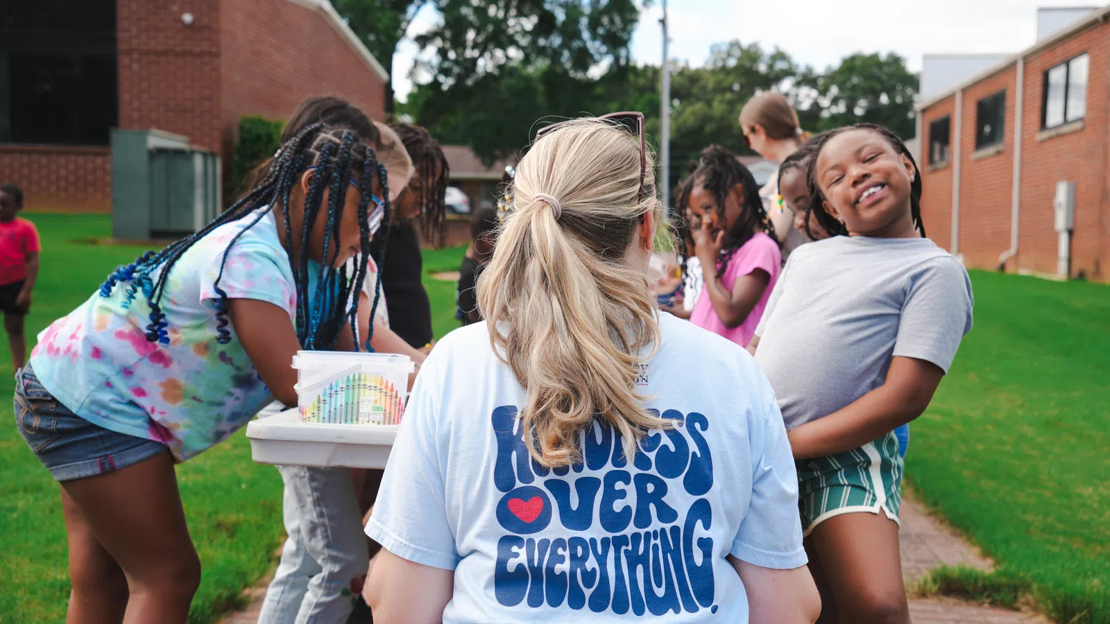 Teacher with kindness t-shirt surrounded by smiling children playing outdoors near school buildings.