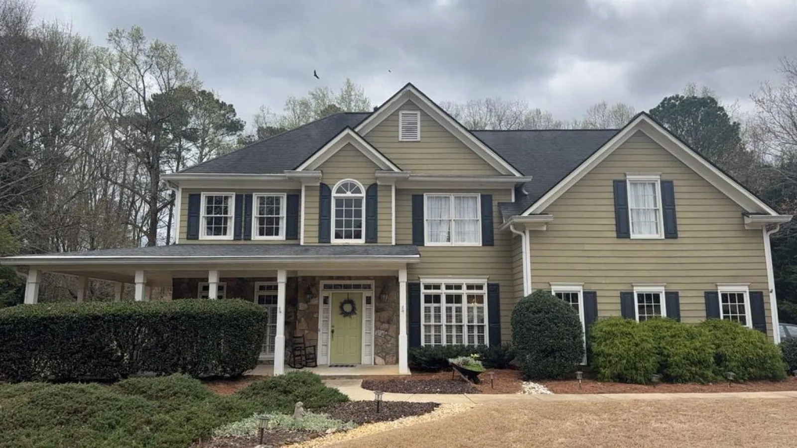Two-story beige suburban house with black shutters, a front porch, manicured bushes, and a green door under a cloudy sky