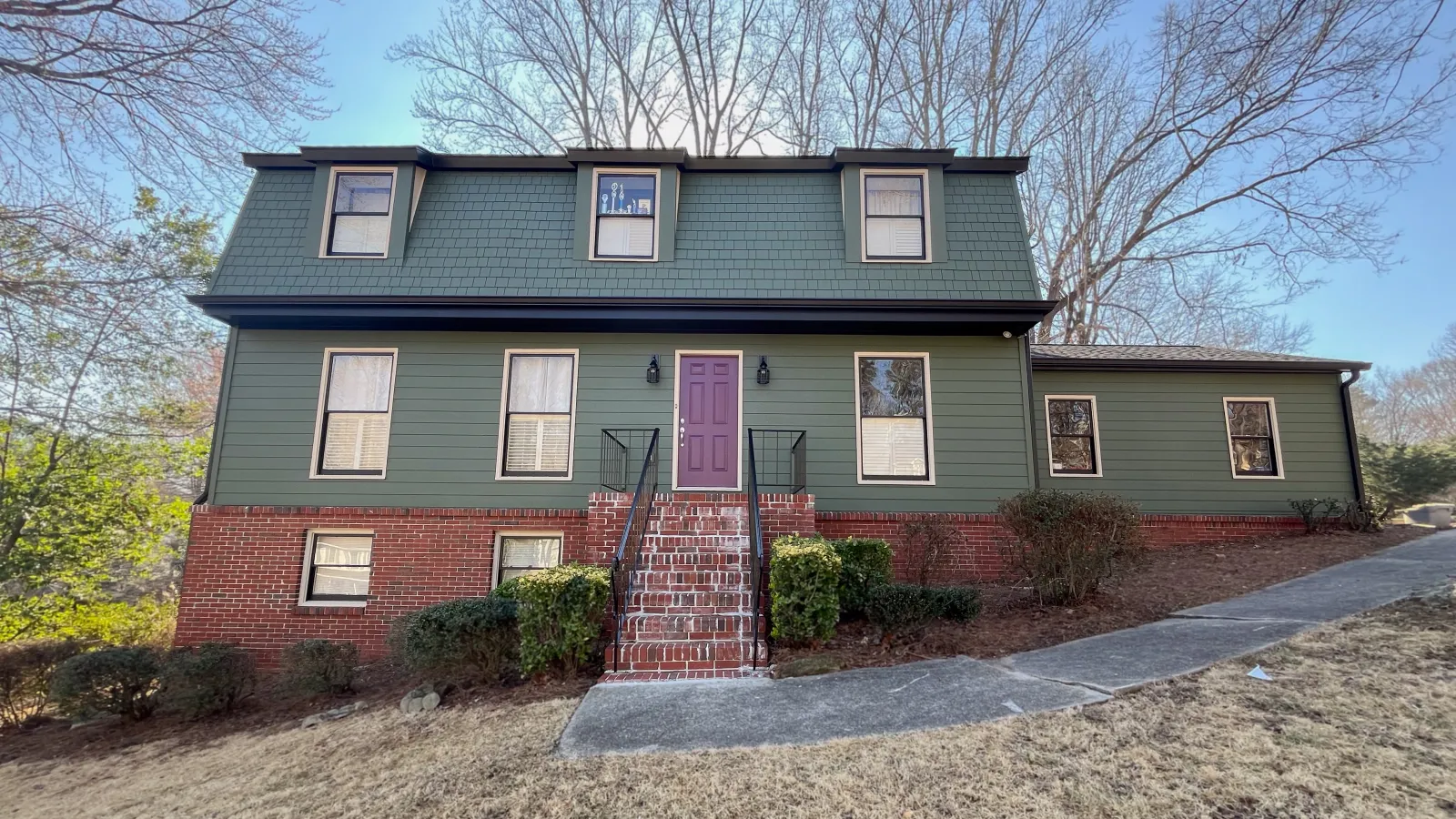 Two-story green house with brick foundation, purple front door, and leafless trees in the background under a blue sky.