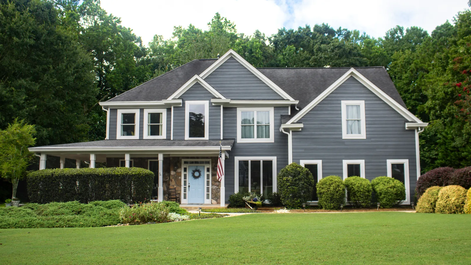 Large two-story blue house with white trim, lush green lawn, and surrounding trees in a suburban setting.