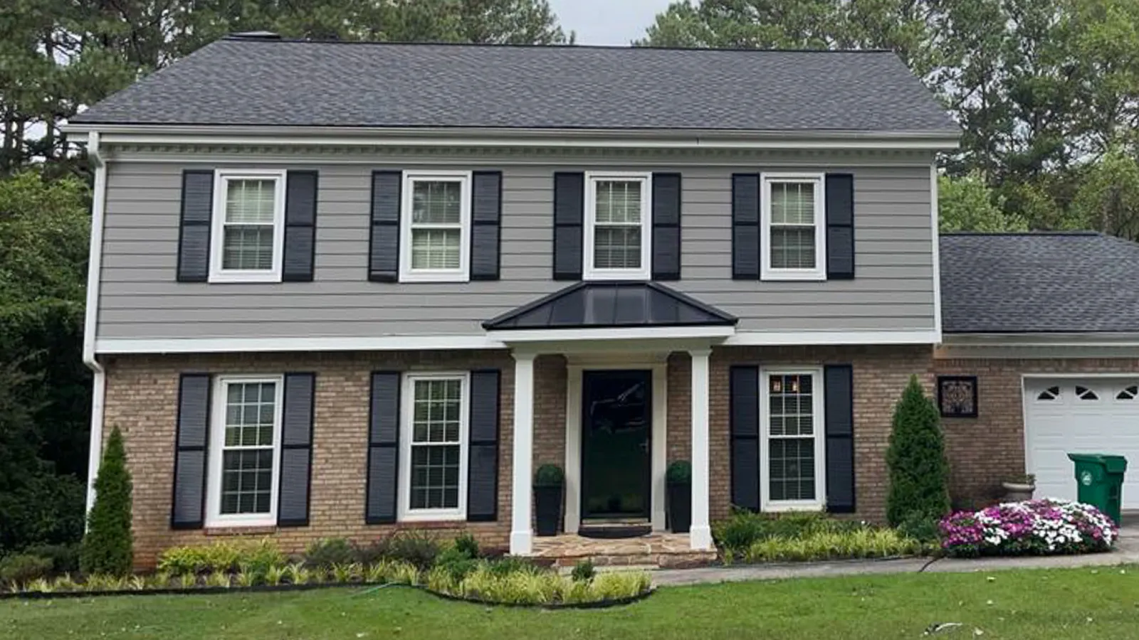 Two-story suburban house with gray siding, brick facade, black shutters, and well-maintained front lawn with shrubs.