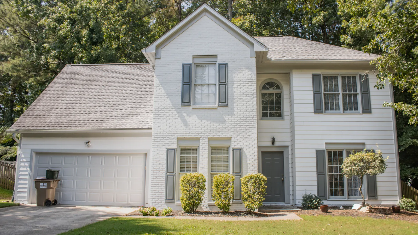 Two-story white house with gray shutters, a double garage, small bushes, and a green lawn under trees.