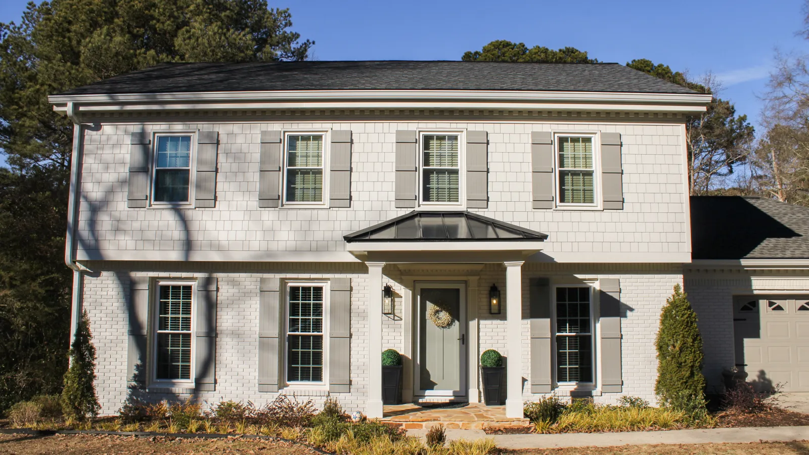 Two-story white brick house with gray shutters, front porch, and landscaped yard under a clear blue sky.