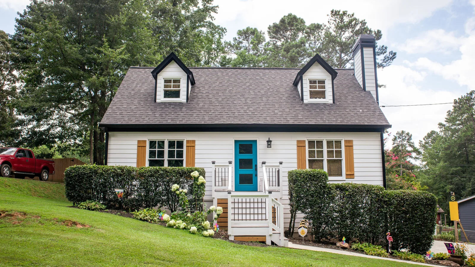 Charming white house with blue door, dormer windows, wooden shutters, and manicured front yard with bushes and flowers.