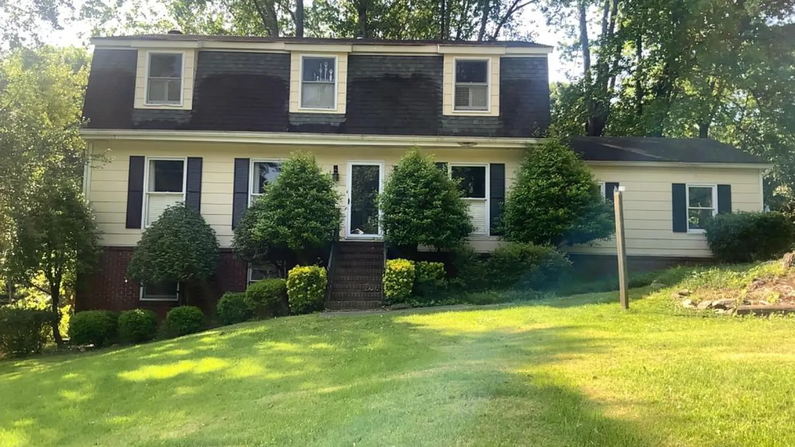 Two-story house with dark roof, cream siding, surrounded by green bushes and trees on a sunny day.