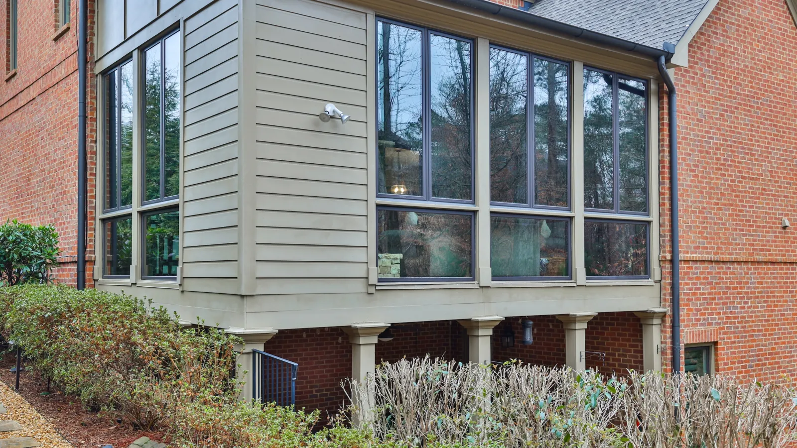 Modern house extension with large glass windows and brick exterior supported by columns above shrubbery.