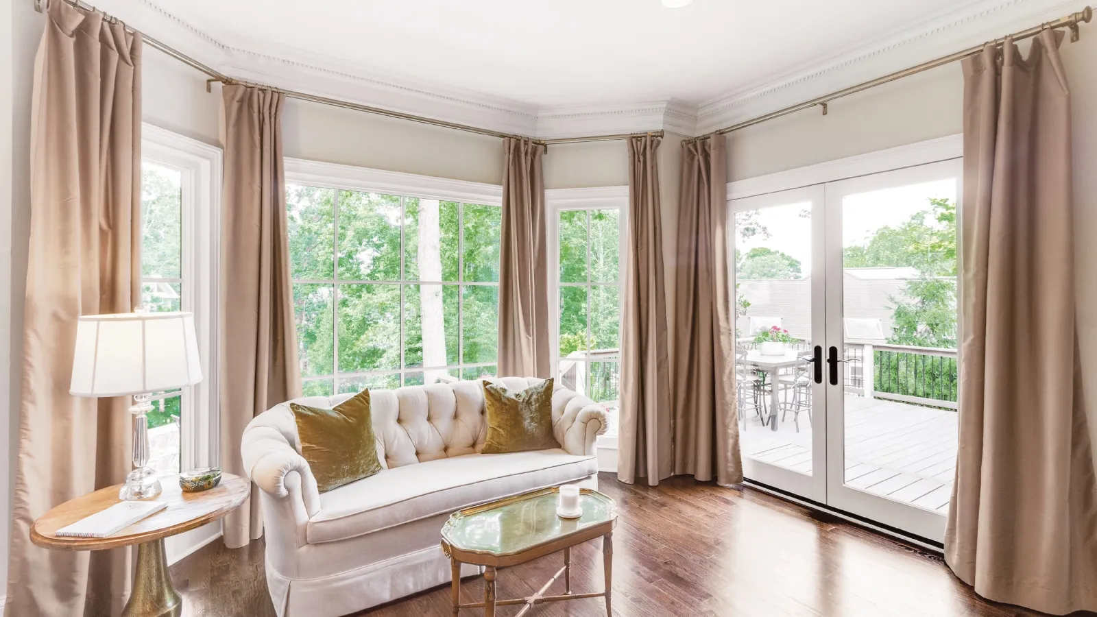 Bright living room with white sofa, beige curtains, glass coffee table, hardwood floor, and large windows with garden view.