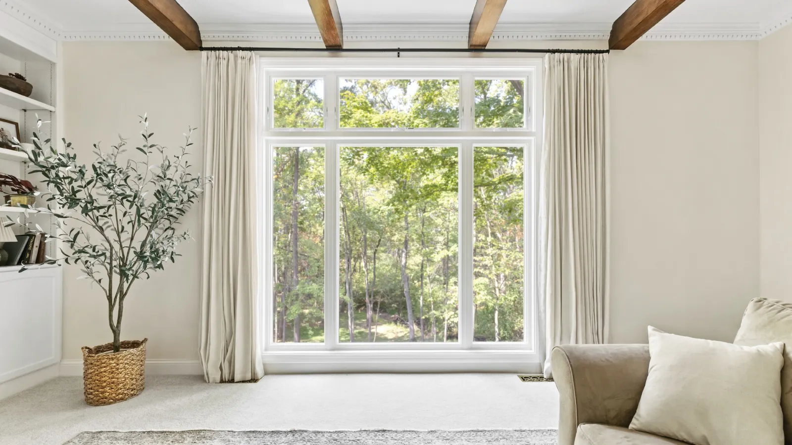 Bright living room with large window, beige sofa, green plant in wicker basket, and wooden ceiling beams.