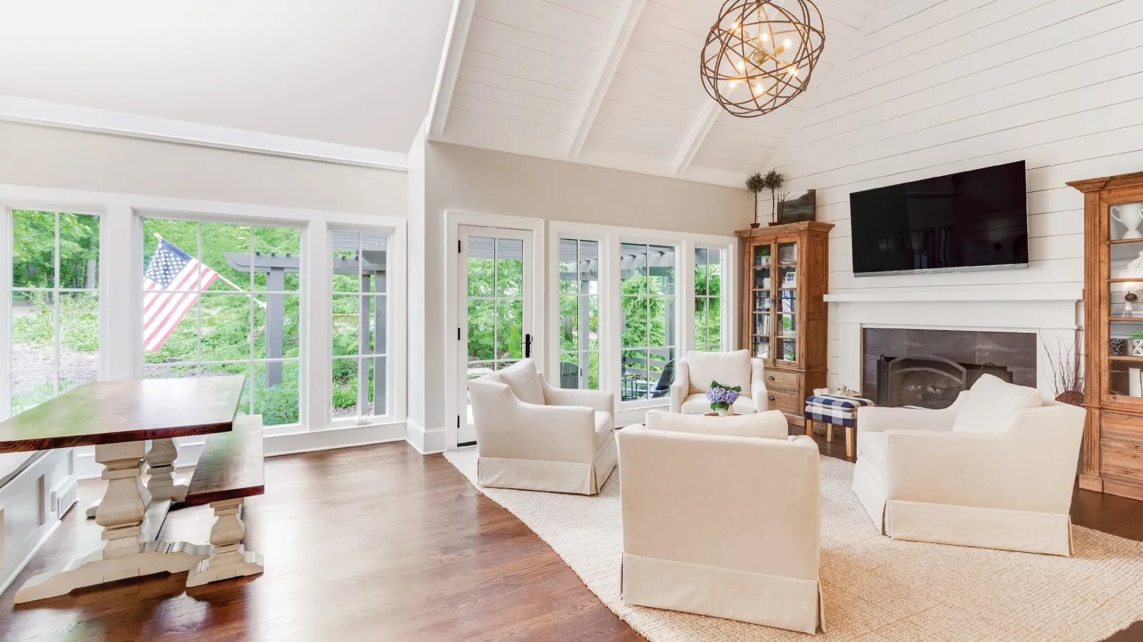 Bright living room with high white ceiling, beige armchairs, wooden flooring, fireplace, and large windows with garden view