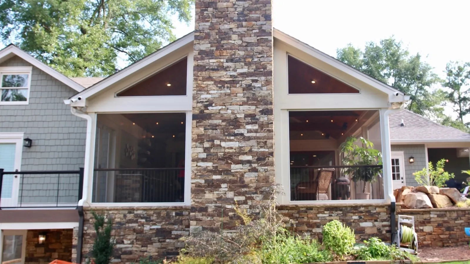 Backyard view of a house with a tall stone chimney, screened porch, lawn, and garden landscape under bright daylight