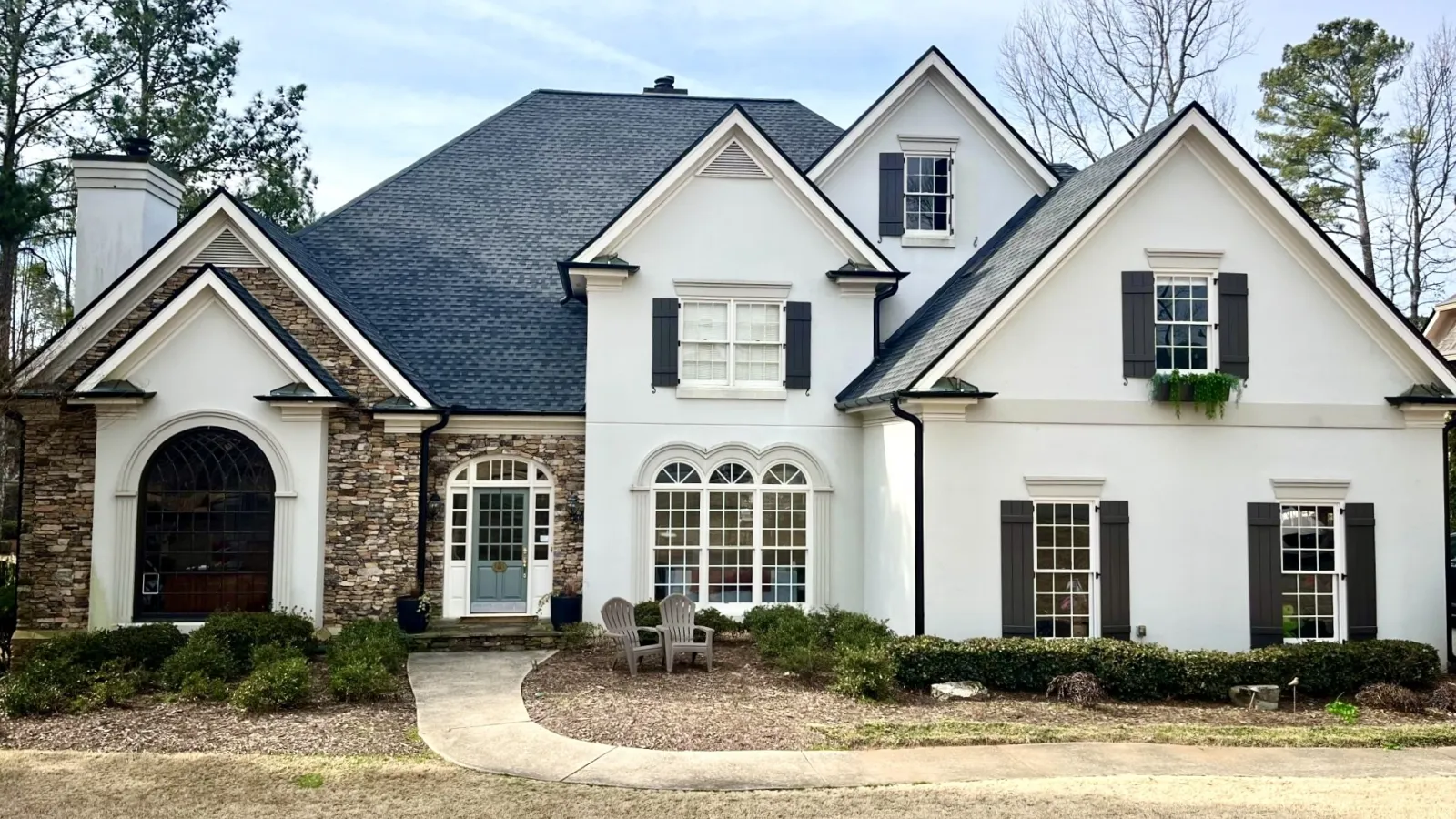 Large white two-story house with black shutters, stone chimney, and front yard with walkway and chairs.