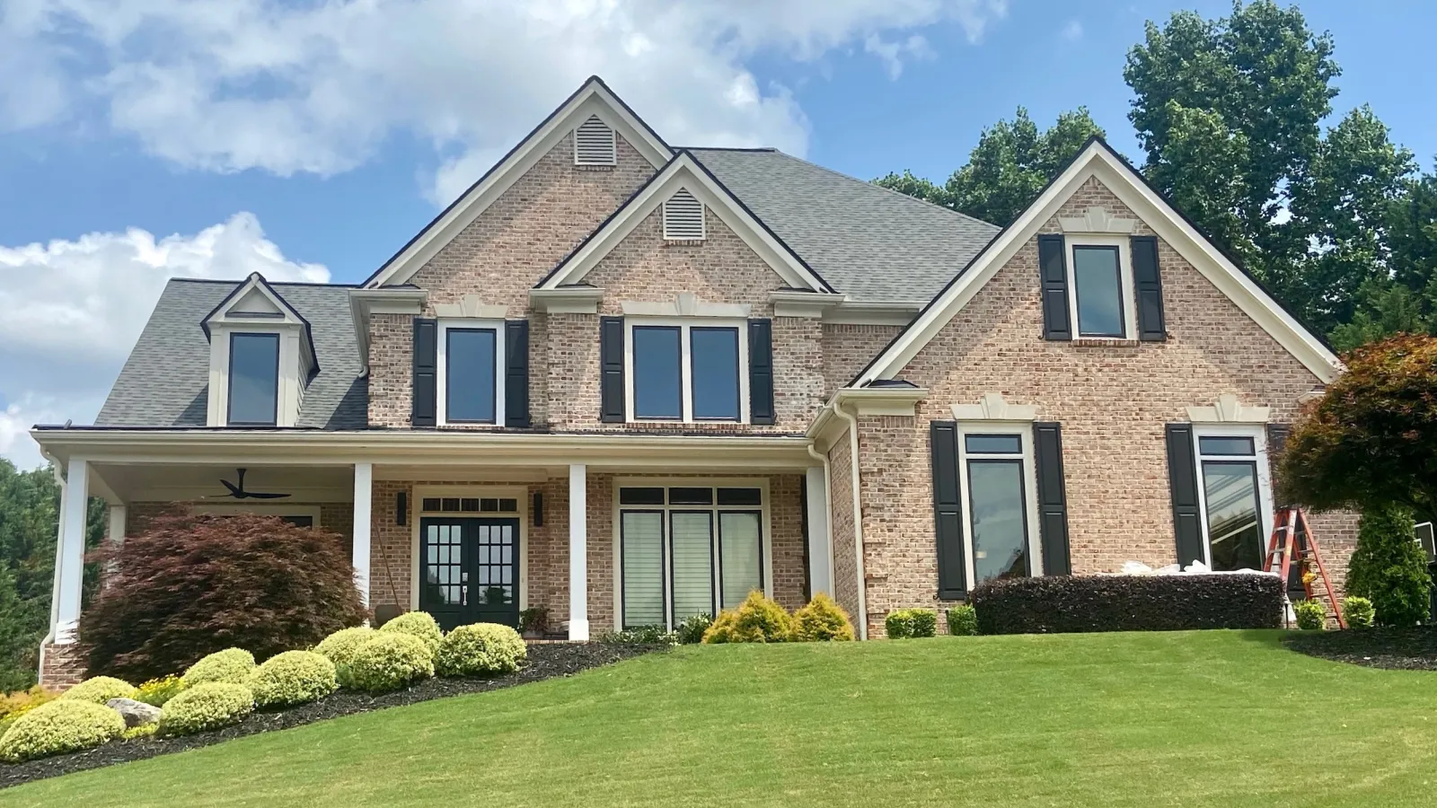Two-story brick house with black window shutters, landscaped front yard, covered porch, and sloped green lawn.