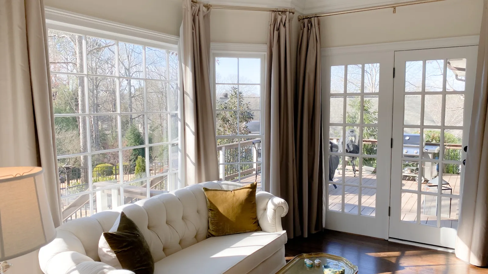 Bright living room with white tufted sofa, large windows, beige curtains, and glass doors leading to a sunny deck.
