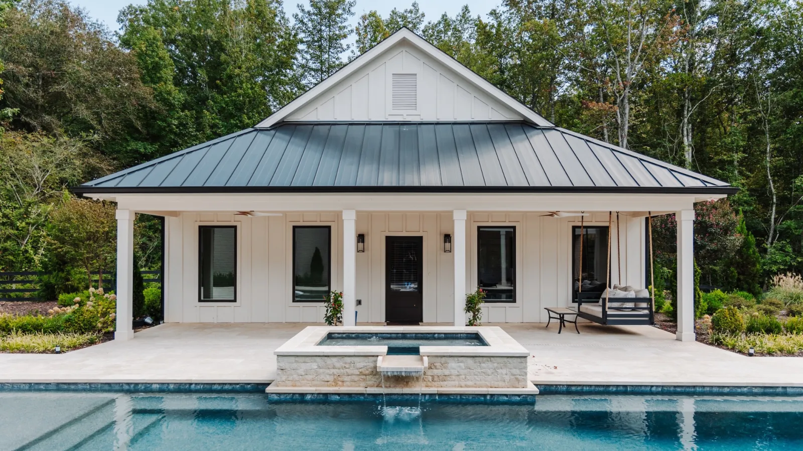 Modern pool house with metal roof, pool, hot tub, and swing surrounded by trees and greenery.